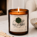 A close-up of a lit hand-poured 11 oz amber jar candle from The Type C Method with a proprietary sage and sandalwood label, resting on a white marble surface next to wooden matches with a blurred fireplace background.