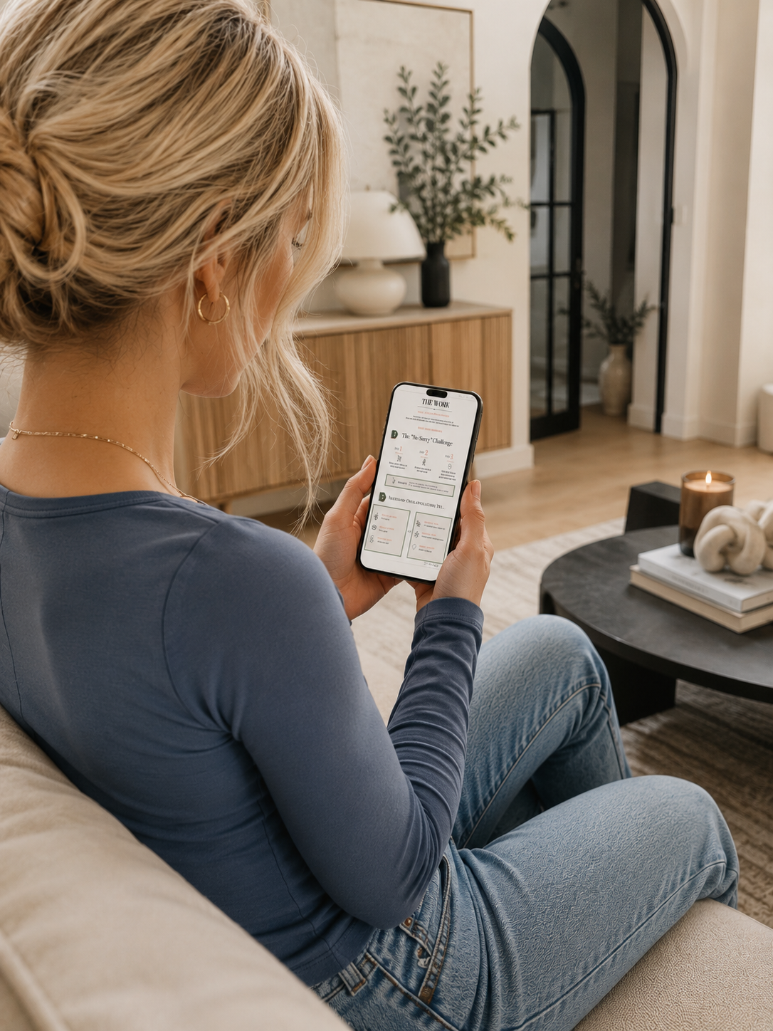Back view of a woman on a cream couch reading the digital Type C Method tracking sheet for the No Sorry Challenge on her smartphone in a modern, neutral-toned living room.