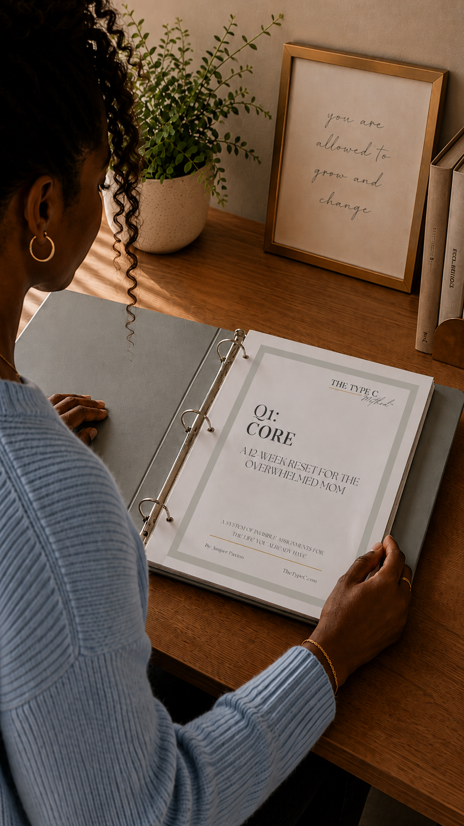 Over-the-shoulder view of a woman at a wooden desk opening a sage green binder containing the printed version of the Type C Method Q1 Core workbook.